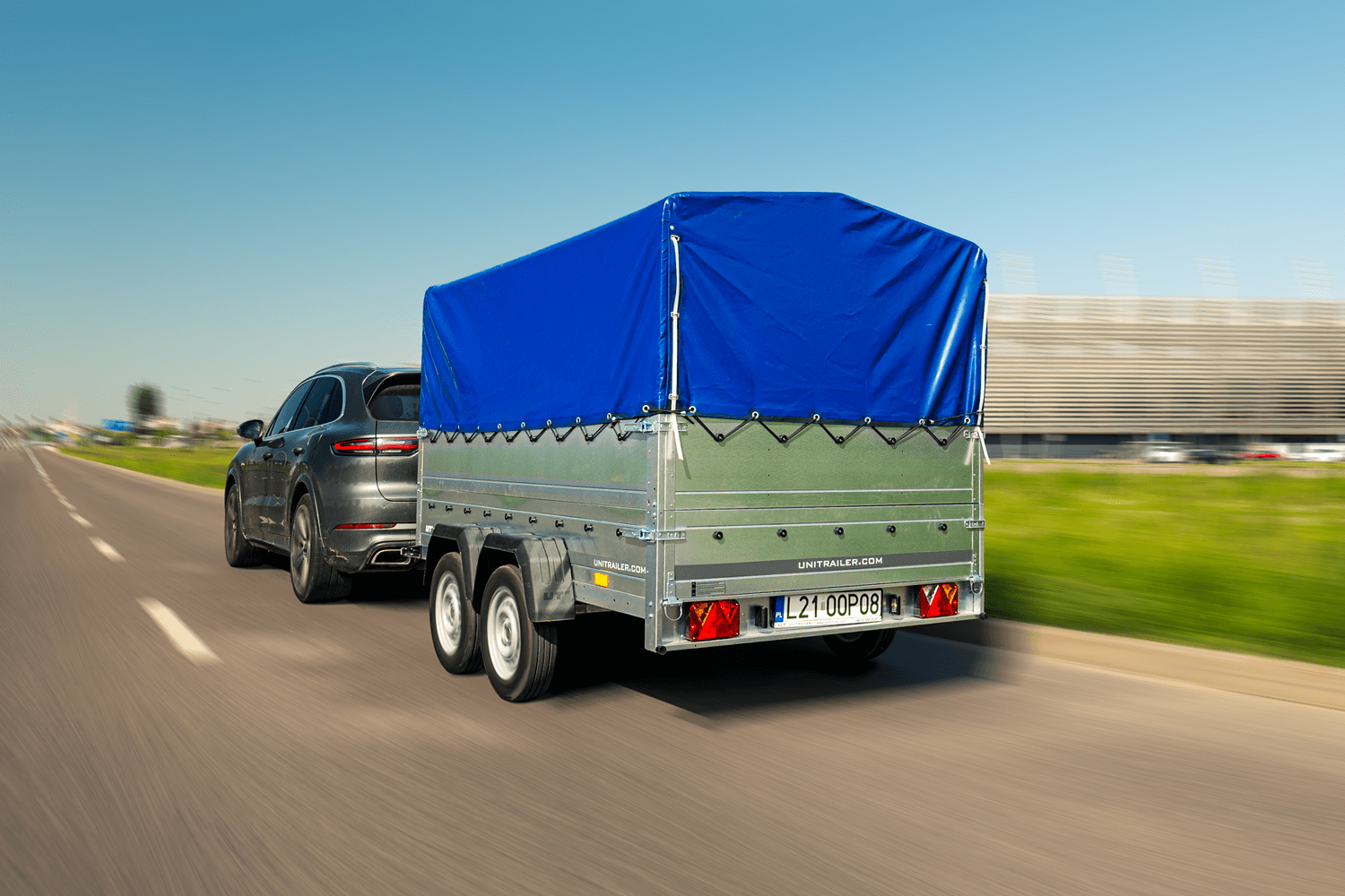 Nowoczesny, ciemnoszary SUV (Porsche Cayenne) porusza się po szerokiej drodze, ciągnąc dużą, dwuosiową przyczepę samochodową Unitrailer. Przyczepa wyposażona jest w wysoką, dokładnie naciągniętą plandekę w kolorze intensywnego błękitu, która jest przymocowana elastyczną linką przeciągniętą przez oczka w burtach. W tle widoczne jest czyste, jasnoniebieskie niebo oraz zarys nowoczesnej bryły architektonicznej. Przyczepa posiada tablicę rejestracyjną L21 00P08.