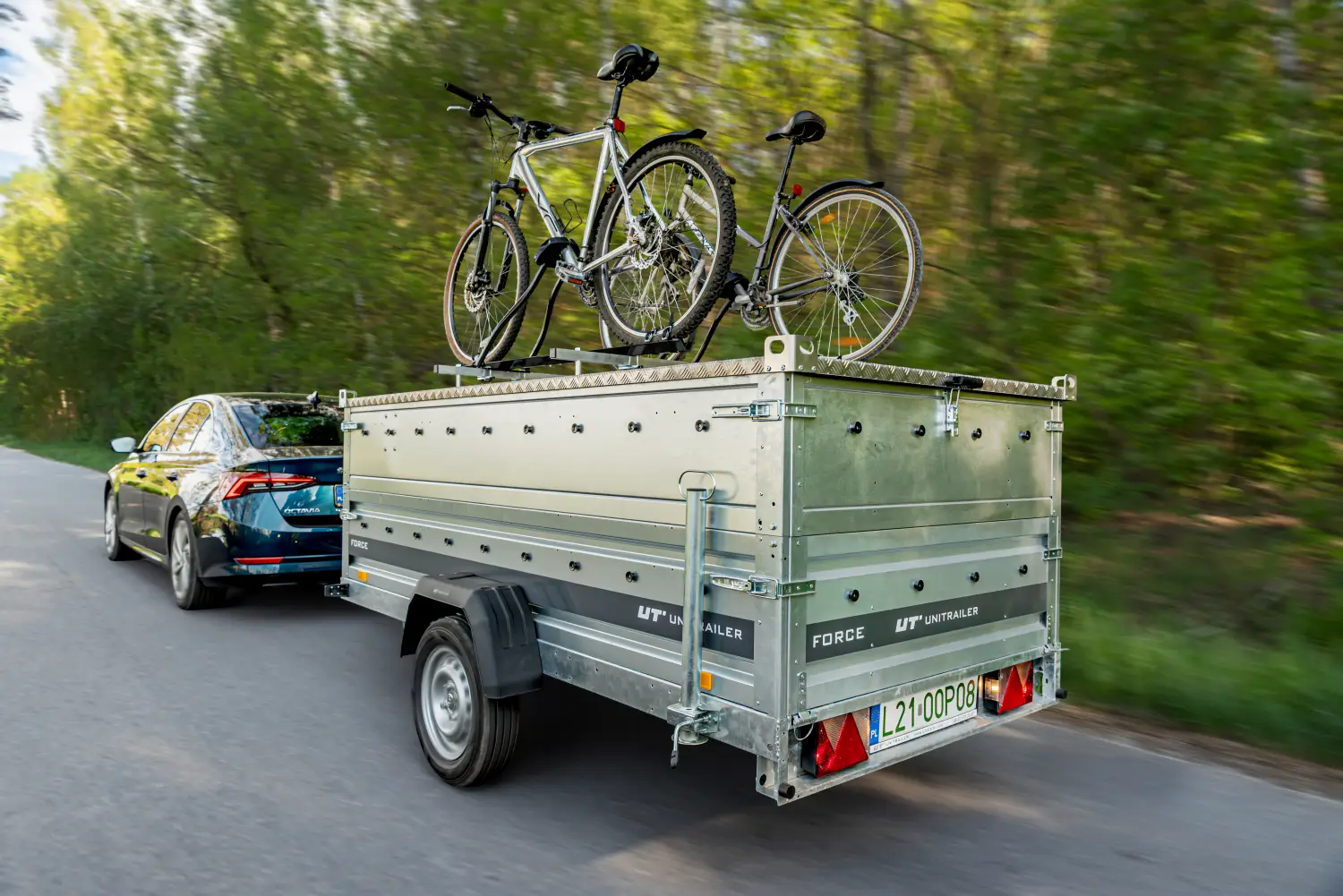 Dynamiczne ujęcie tyłu ciemnoniebieskiego samochodu osobowego (Skoda Octavia) jadącego asfaltową drogą i ciągnącego srebrną przyczepę jednoosiową marki Unitrailer Force. Przyczepka samochodowa wyposażona jest w potrójne, wysokie burty (nadstawki) oraz aluminiową, ryflowaną pokrywę burtową. Na pokrywie zamontowano dwa rowery – srebrny górski oraz ciemny rower trekkingowy. W tle widoczne są rozmyte zielone drzewa w słoneczny dzień. Na tylnej burcie znajduje się tablica rejestracyjna L21 00P08 oraz trójkątne odblaski.
