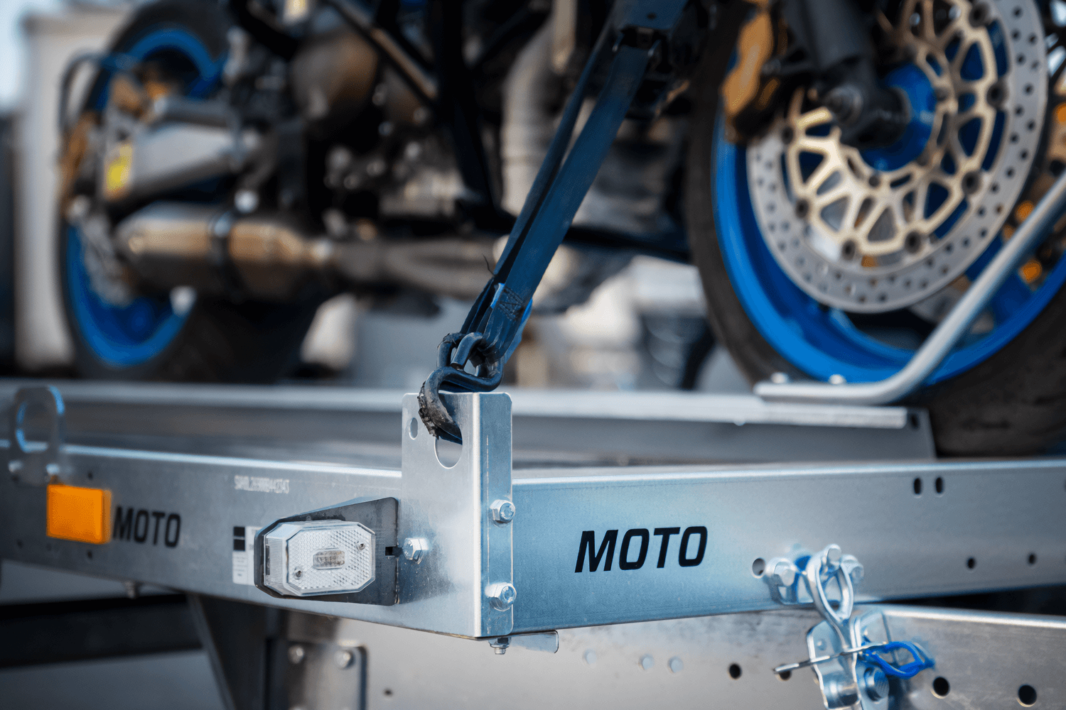 Bliskie ujęcie detali przedniego koła motocykla zabezpieczonego pasami transportowymi na przyczepie motocyklowej. Czarny, solidny pas transportowy z hakiem jest napięty i zamocowany do specjalnego uchwytu w bocznej, ocynkowanej rynnie z wyraźnym czarnym napisem „MOTO”. W kadrze widoczna jest sportowa, niebieska felga motocykla, perforowana tarcza hamulcowa oraz złoty zacisk hamulcowy, co podkreśla profesjonalne przygotowanie jednośladu do transportu.