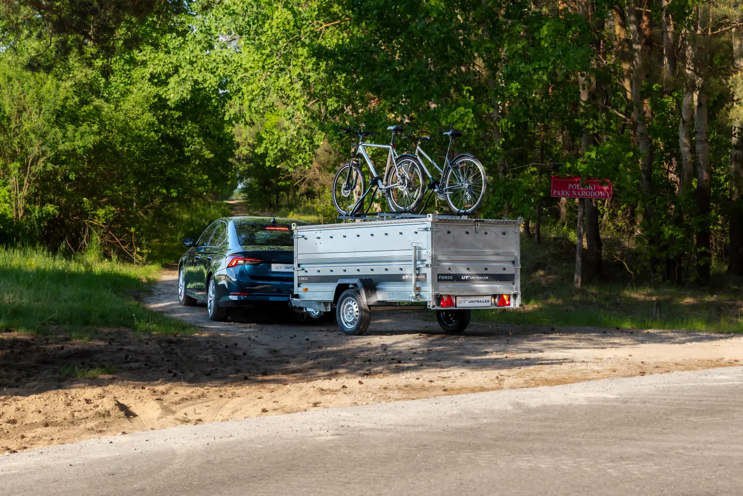 Ciemnoniebieski sedan zjeżdżający na piaszczystą leśną ścieżkę obok czerwonej tablicy z napisem „POLESKI PARK NARODOWY”. Samochód ciągnie przyczepę z podwyższoną aluminiową pokrywą, na której zainstalowano uchwyt z dwoma rowerami.