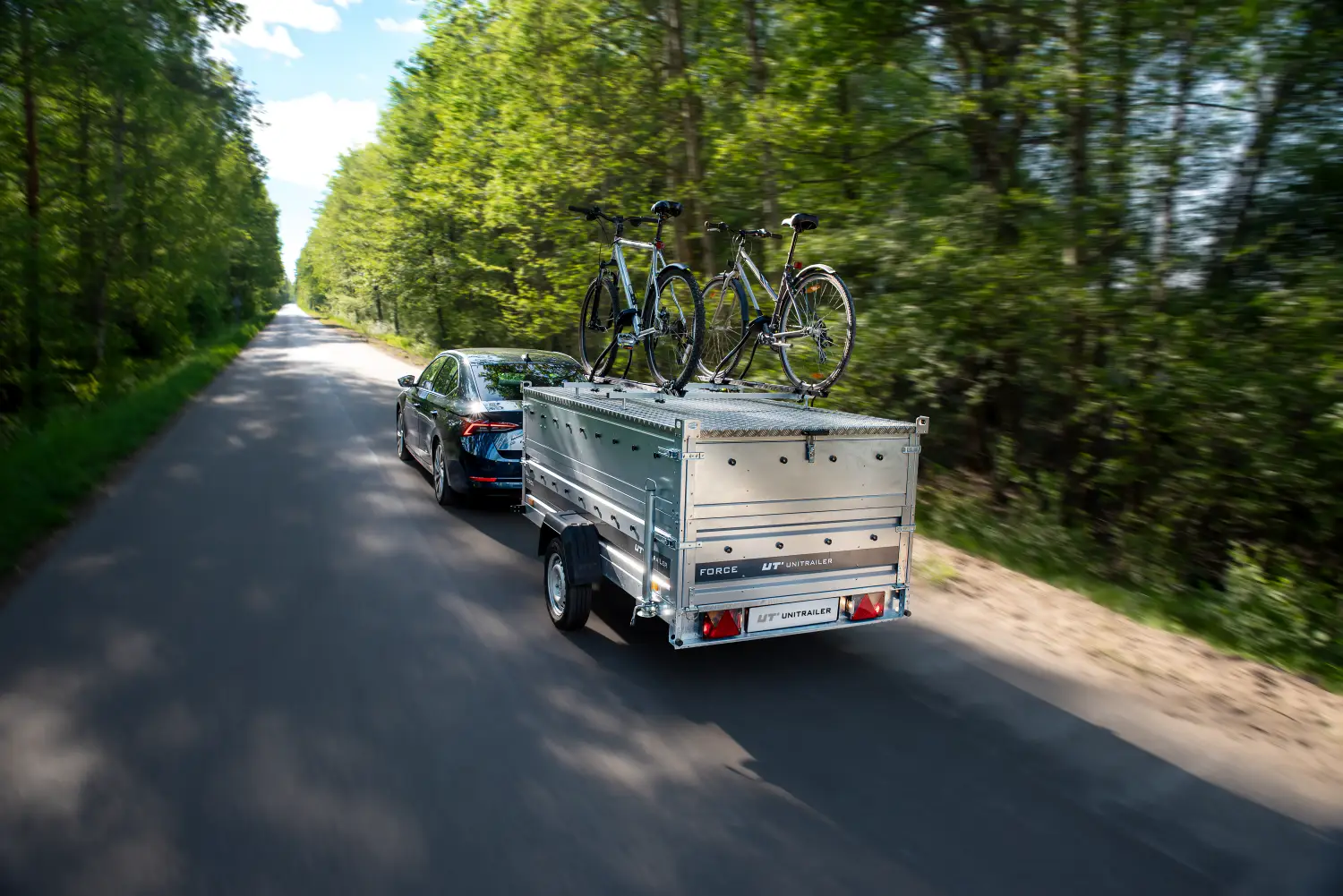 Ciemnoniebieski sedan jadący asfaltową drogą przez las, ciągnący przyczepę Unitrailer z sztywną pokrywą aluminiową oraz zamontowanym uchwytem z dwoma rowerami.