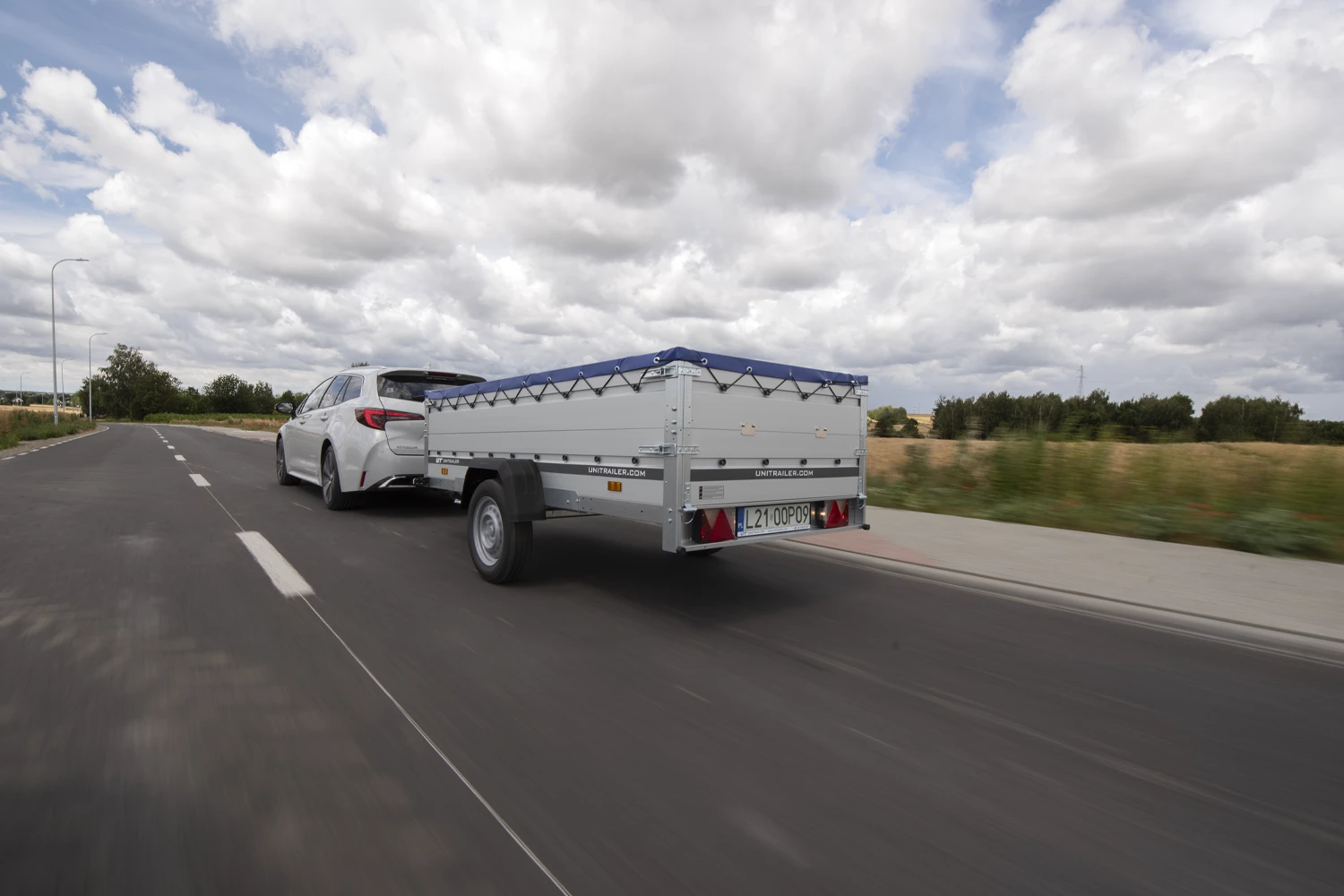 Dynamiczne zdjęcie w ruchu (wykonane techniką panoramowania) przedstawiające srebrny samochód Toyota kombi jadący drogą asfaltową i ciągnący za sobą srebrną przyczepę Unitrailer z niebieską plandeką. Pojazd porusza się w prawą stronę kadru wzdłuż białej linii ciągłej oddzielającej pas ruchu od pobocza. Całe tło za samochodem – obejmujące zielone zarośla, drzewa oraz odległe pola – jest mocno rozmyte, co skutecznie oddaje wrażenie prędkości. Nad horyzontem rozciąga się dynamiczne, pełne ekspresji niebo z dużymi, kłębiastymi biało-szarymi chmurami.