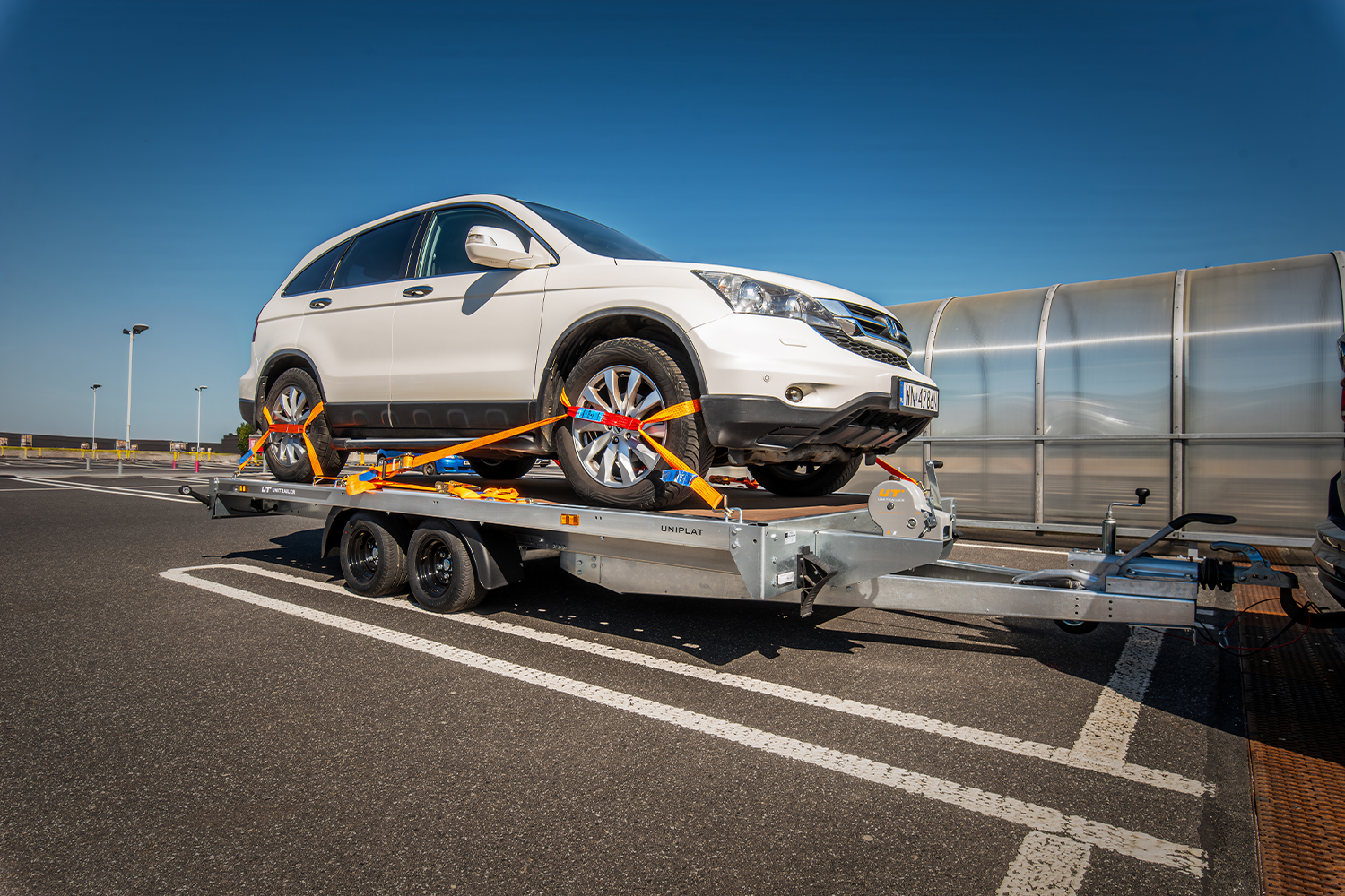 Widok z poziomu asfaltu. Parking z białymi liniami. W tle wiata na wózki zakupowe oraz latarnie. Czyste, jasne, niebieskie niebo. W kadrze przyczepa w typie lawety z napisem 