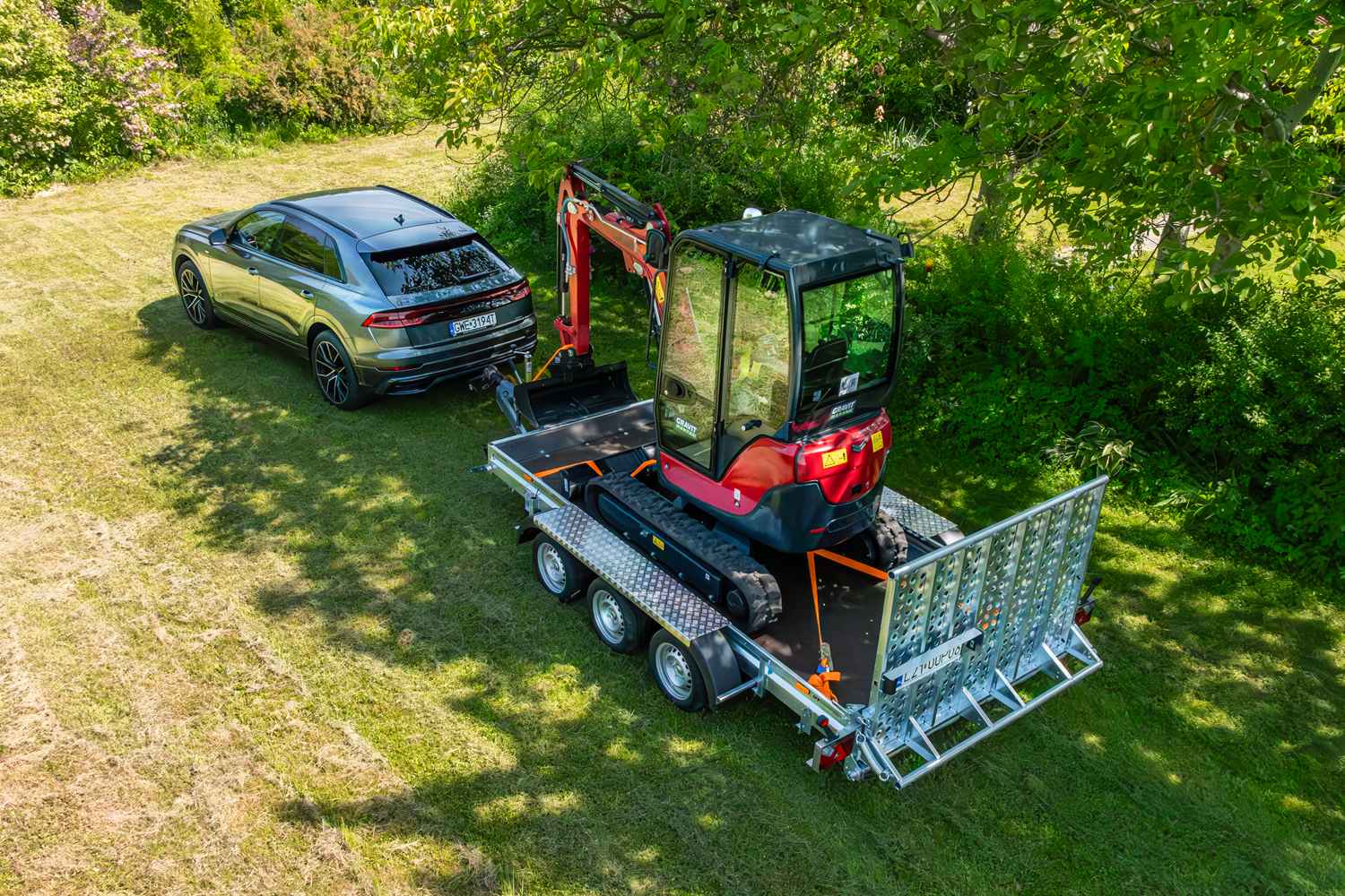 Widok z góry na niewielką polanę, otoczoną drzewami. Słoneczny dzień. Na polanie srebrne, nowoczesne auta z zaczepioną do niego lawetą trzyosiową, na której zamontowana jest mini koparka w kolorze czerwonym.