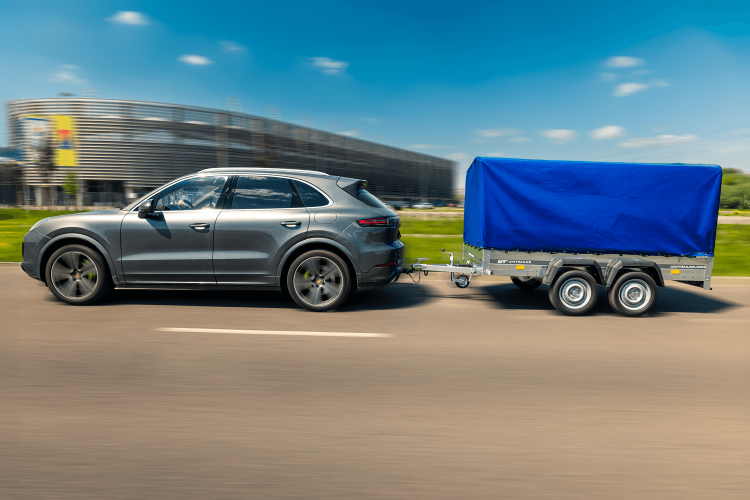 Widok od boku na samochód jadący z przyczepą. W tle stadion piłkarski oraz pas zieleni. Samochód typu premium SUV marki Porsche. Przyczepa z logo 