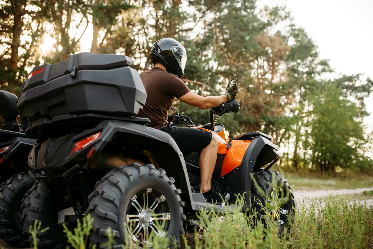 Mężczyzna w kasku motocyklowym siedzi na pomarańczowym quadzie wyposażonym w duży czarny kuferek bagażowy na tle lasu.