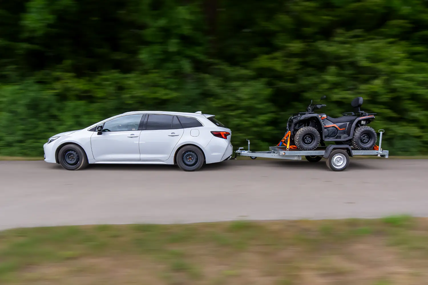 Srebrny samochód kombi w ruchu, holujący na platformie przyczepy transportowej czarnego quada zabezpieczonego pasami.