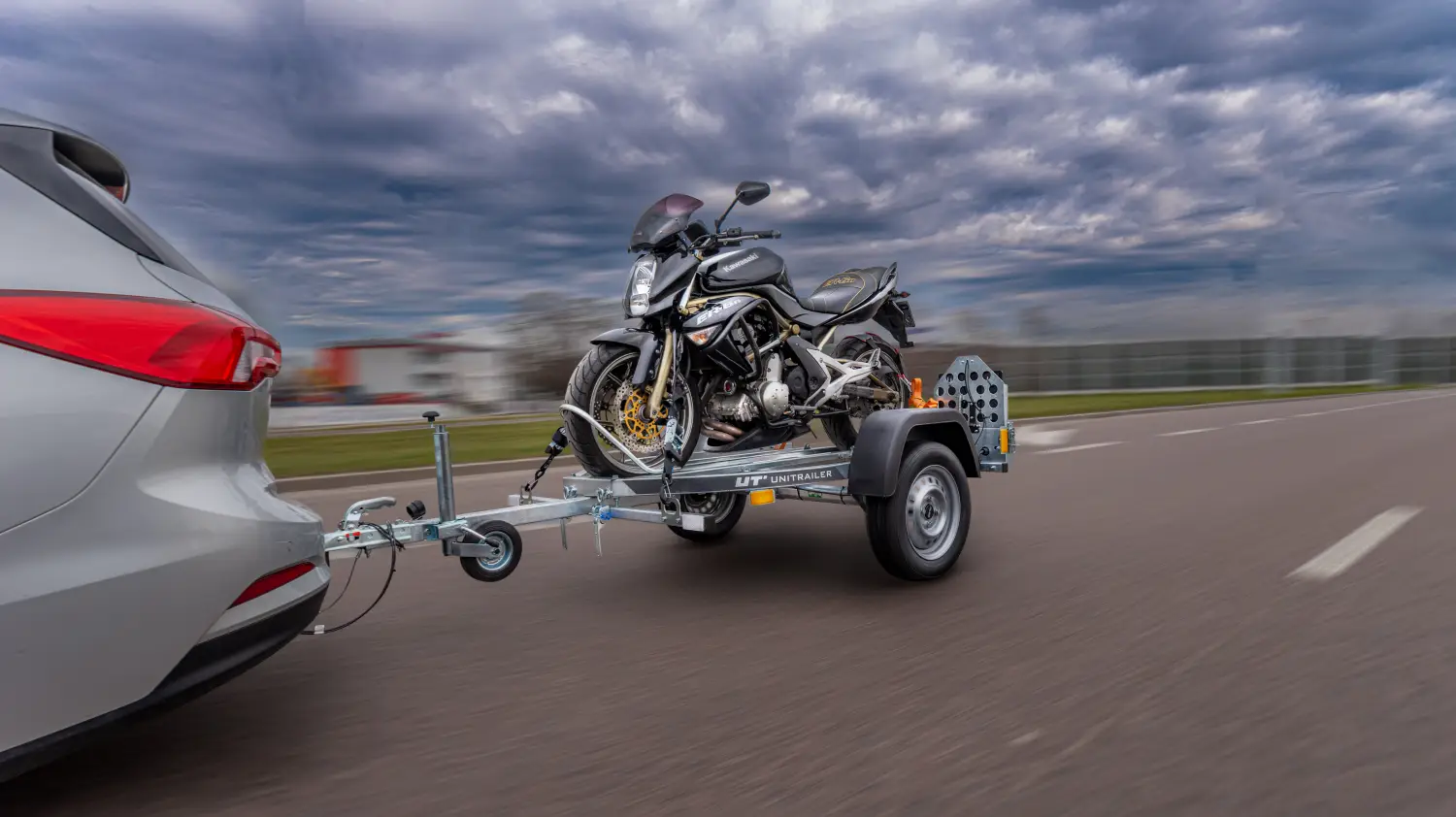 Srebrny samochód osobowy ciągnący jednoolową przyczepę motocyklową marki Unitrailer po drodze asfaltowej. Na przyczepie zamontowany i zabezpieczony pasami jest czarny motocykl szosowy Kawasaki.