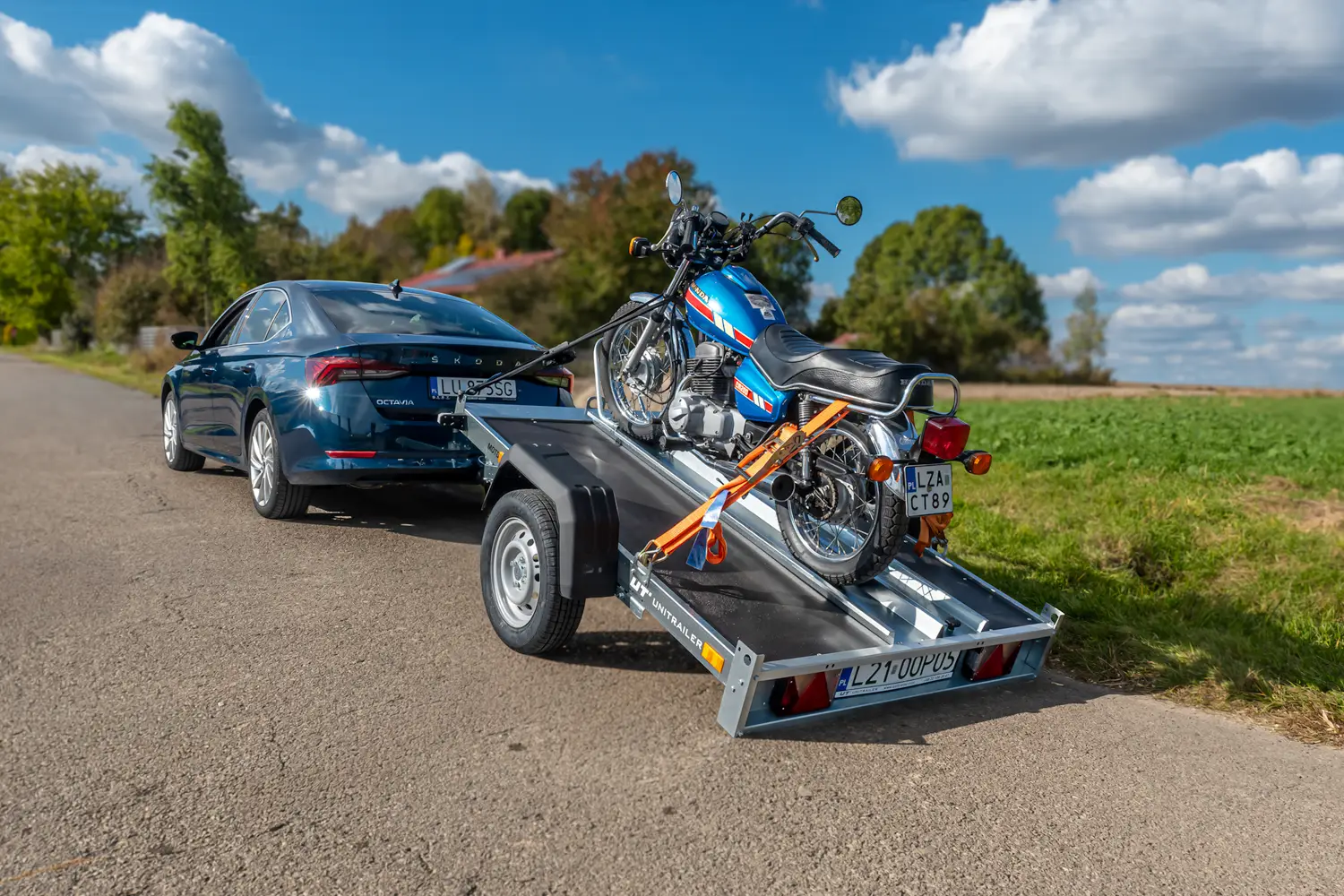 Ciemnoniebieska Škoda Octavia zaparkowana na wiejskiej drodze z uchylną przyczepą motocyklową Unitrailer. Na opuszczonej platformie przyczepy zabezpieczono pasami klasyczny, niebieski motocykl Honda.