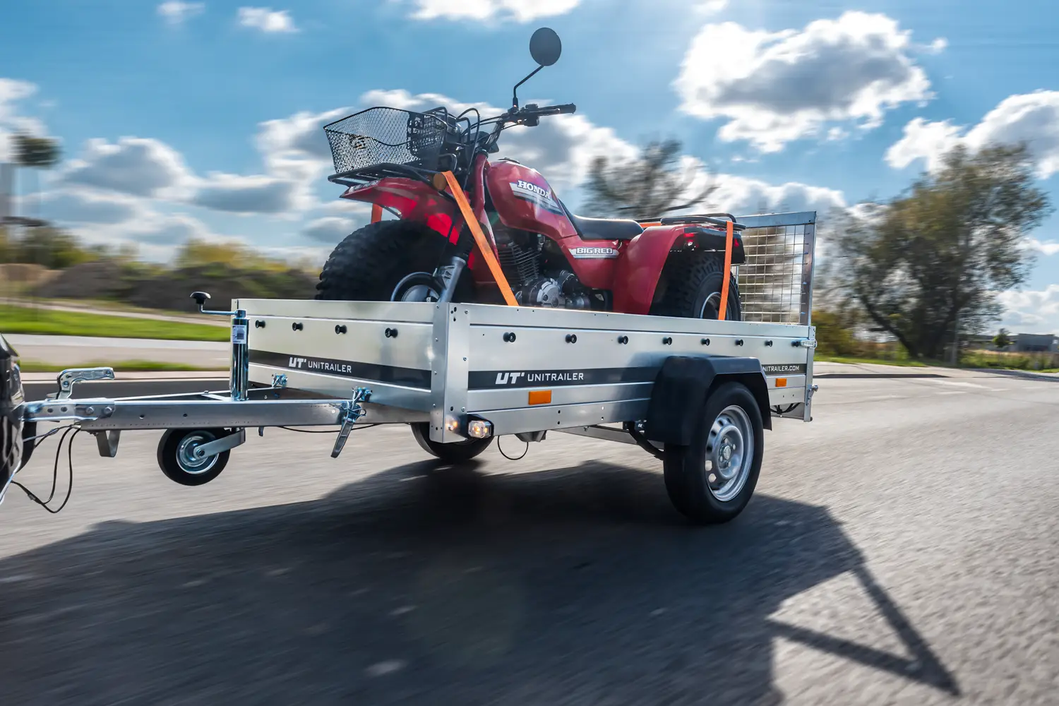 Jednoosiowa przyczepa burtowa marki Unitrailer w ruchu, transportująca czerwony trójkołowiec terenowy Honda z koszykiem na kierownicy, przypięty pomarańczowymi pasami mocującymi.