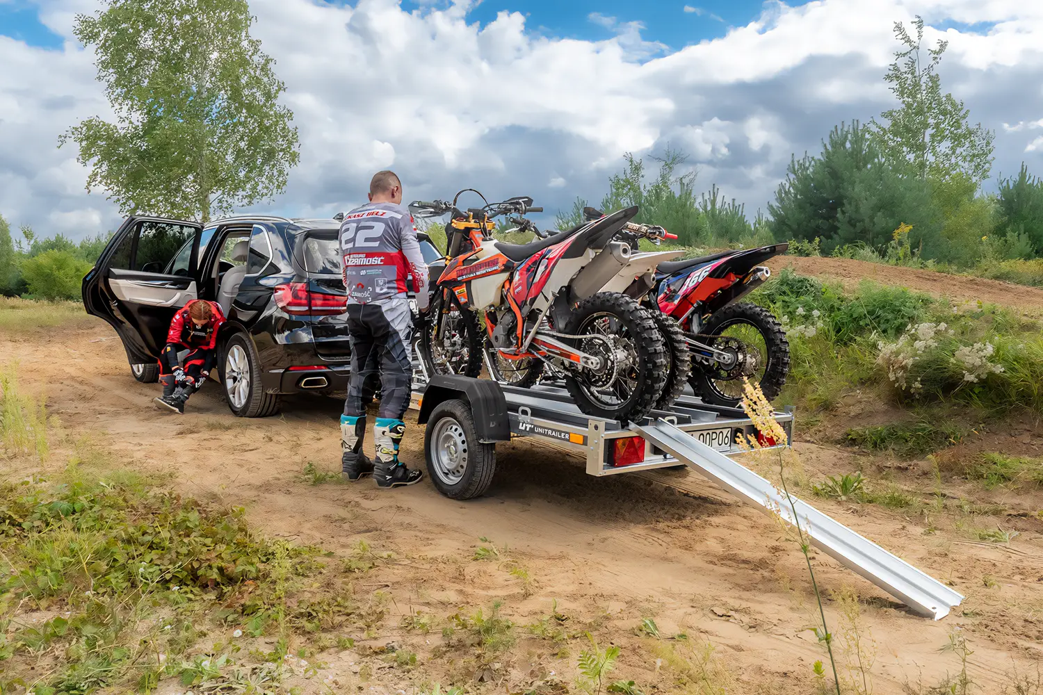 Czarny SUV zaparkowany na piaszczystym torze off-road z przyczepą motocyklową Unitrailer, na której załadowano dwa motocykle crossowe. Dwoje motocyklistów w pełnym stroju ochronnym przygotowuje się do jazdy obok rozłożonej rampy najazdowej.
