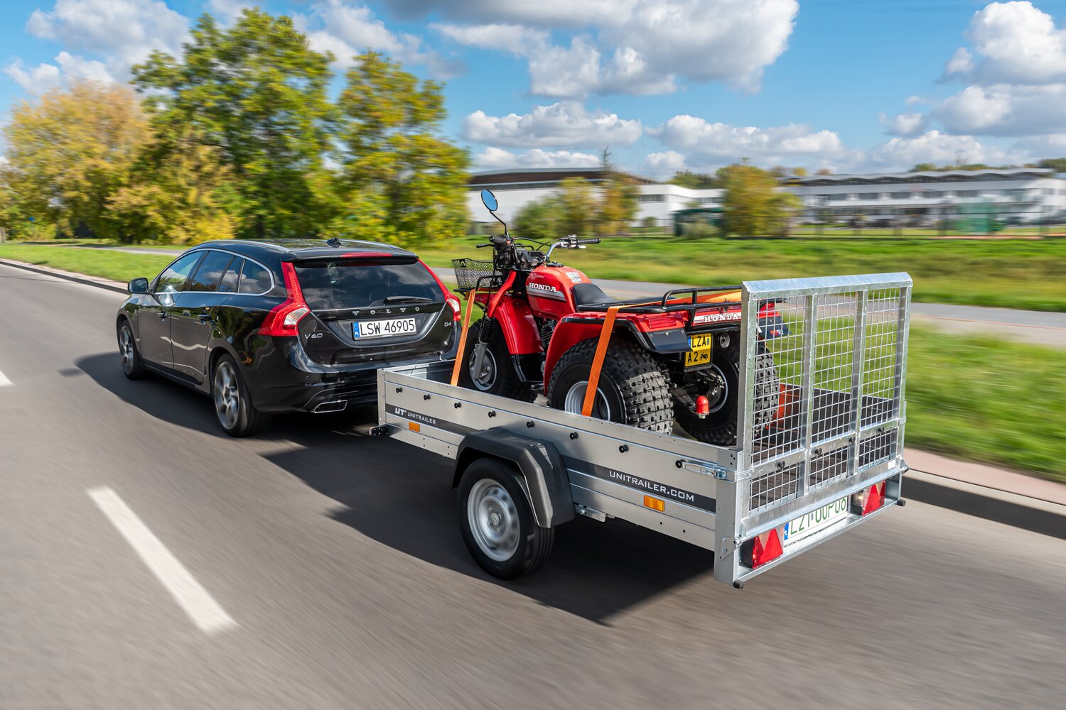 Czarne Volvo holujące srebrną przyczepę samochodową, na której umieszczony jest czerwony quad, zabezpieczony pomarańczowymi pasami transportowymi. W tle tereny zielone oraz zabudowania