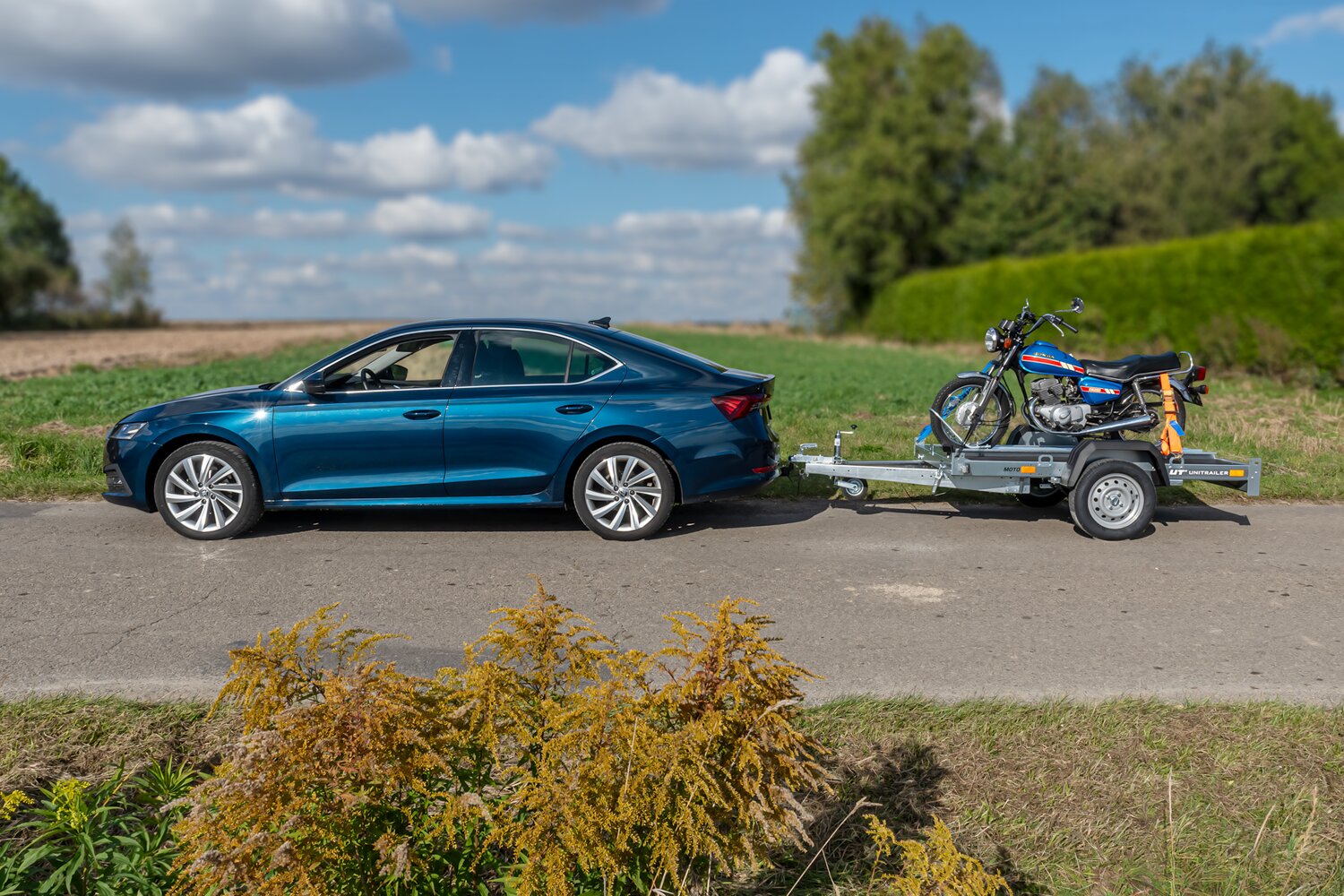 zielona skoda stojąca na asfalcie. W tle zielone pole. Do Skody podczepiona przyczepka MOTO 1 z zmontowanym na niej niewielkim motocyklem jednośladowym typu chopper
