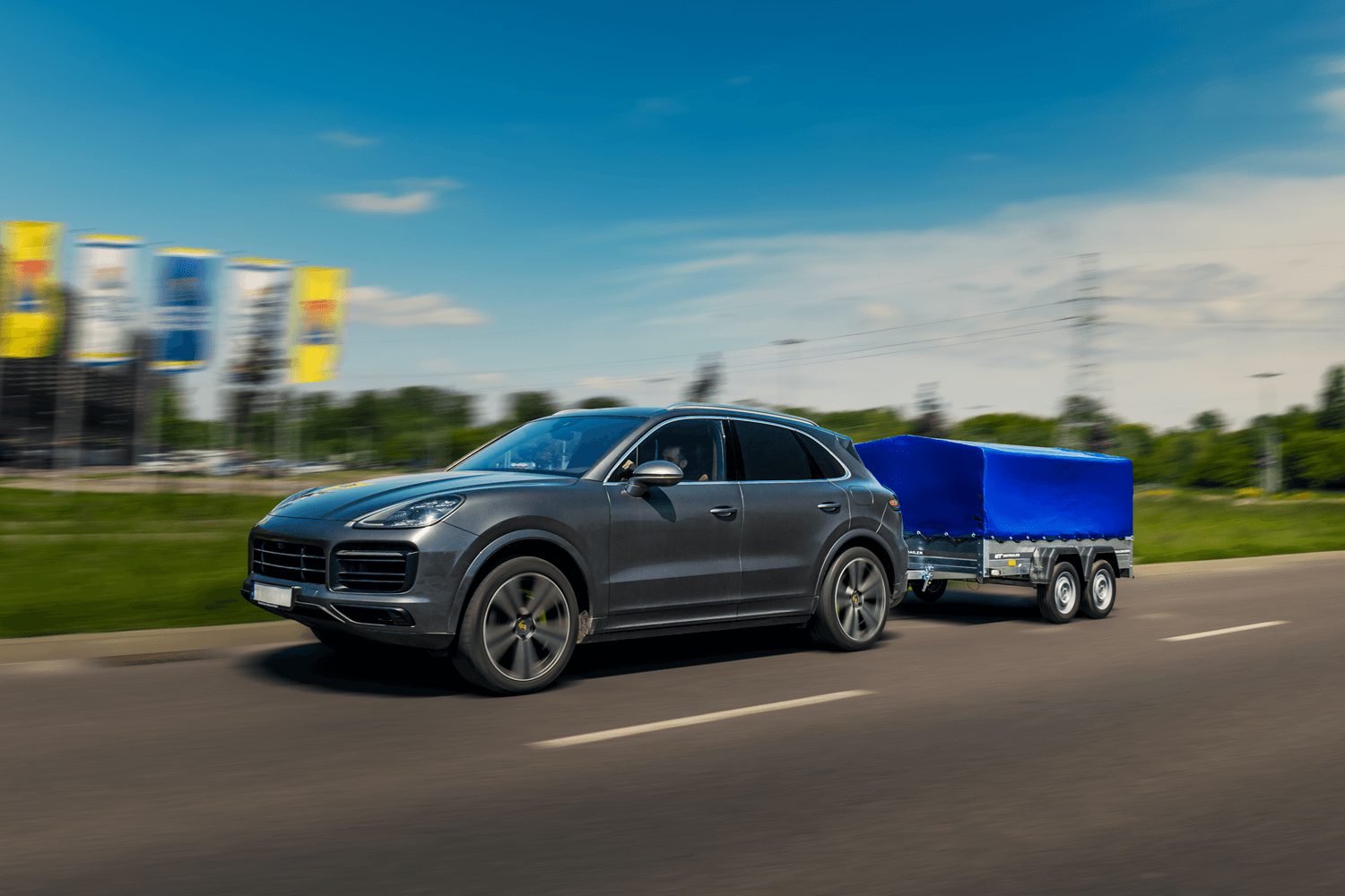Porsche Cayenne w kolorze srebrnym holujące srebrną przyczepkę Unitrailer z zamontowanym stelażem w kolorze niebieskim. Rozmazane tło, na którym widoczny jest las oraz parking z żółtymi i białymi flagami.