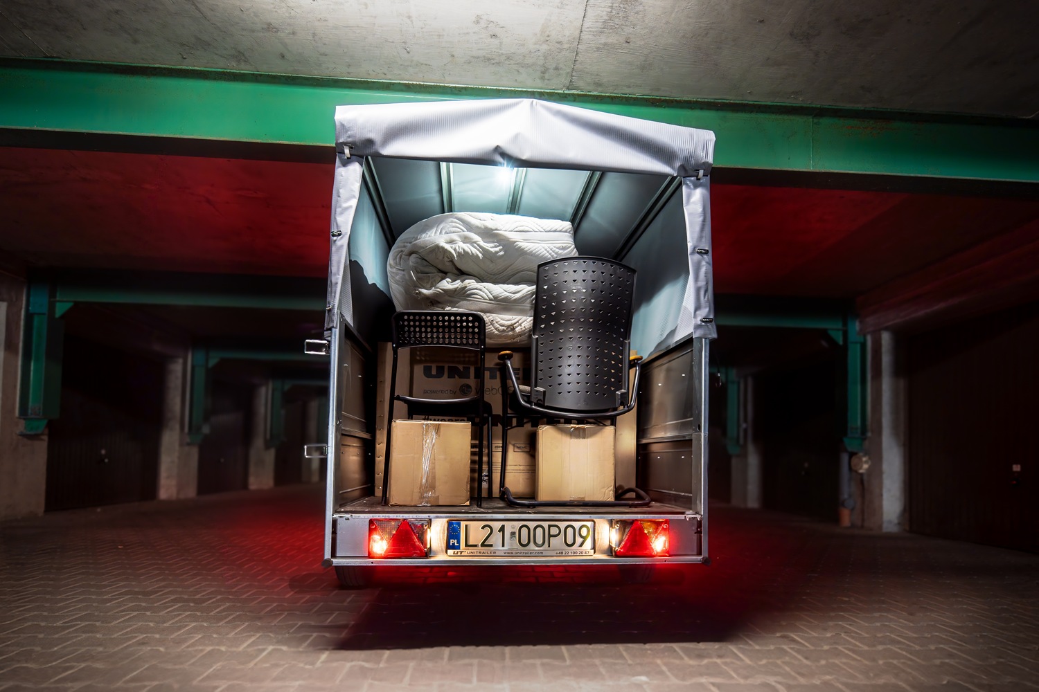 Przyczepa w zaciemnionym garażu. Przyczepa z burtami bis i stelażem szarym. W środku krzesła, pudła i materac. Przyczepa dobrze oświetlona ze źródłem światła wewnątrz konstrukcji oraz na światłach tylnych. Światło wewnątrz białe i jasne.