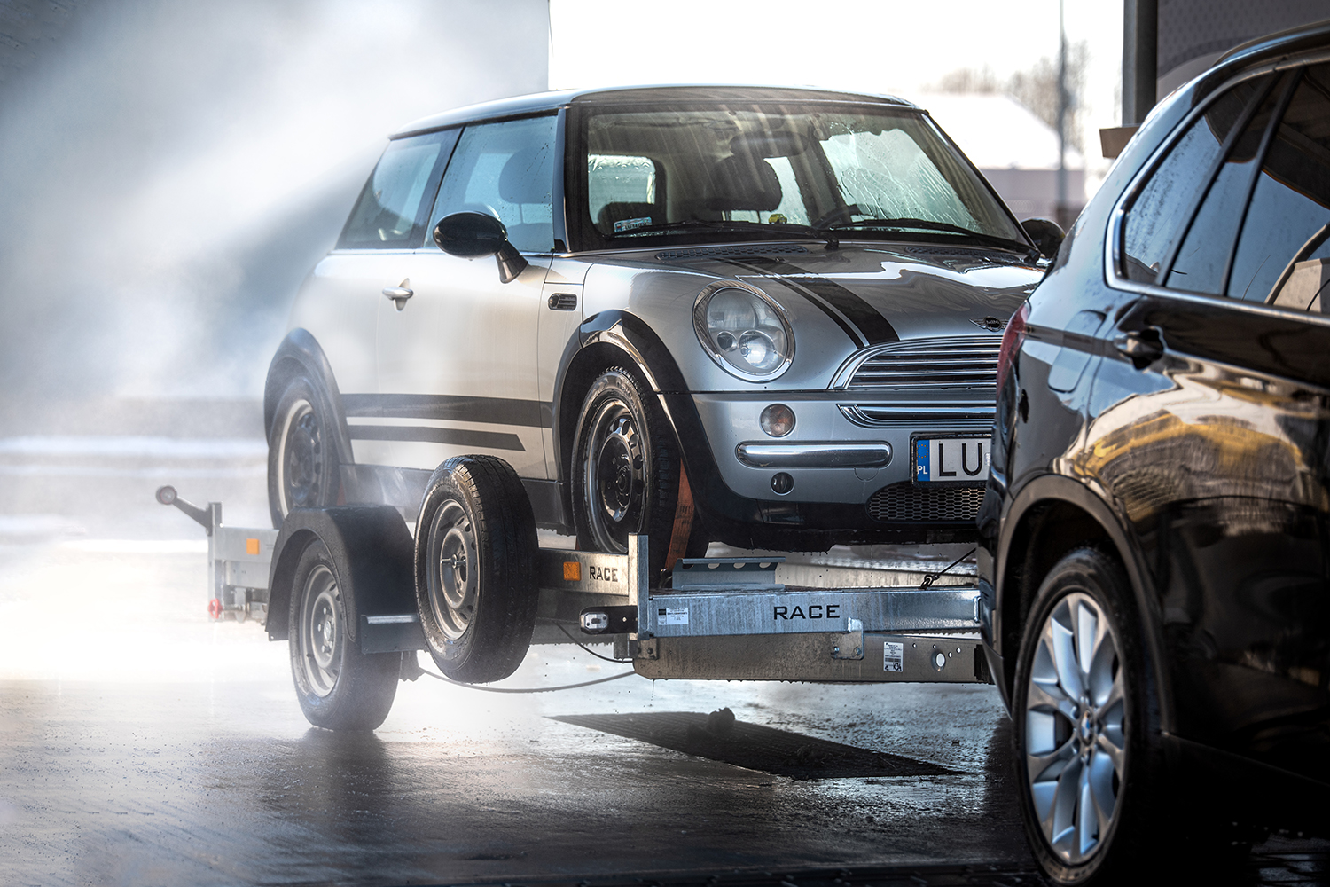 Srebrny samochód marki Mini zamontowany na srebrnej platformie, która holowana jest przez czarne auto. Miejsce: myjnia samoobsługowa. w tle mgiełka z wody i widoczna ścianka myjni.