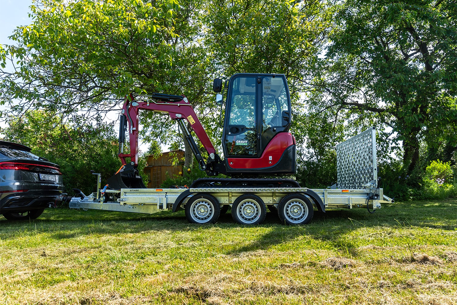 Czerwona mini koparka zamontowana na przyczepie samochodowej. Przyczepa podczepiona do srebrnego SUVa. Teren zielony, przypominający działkę. Trawnik oraz drzewa. Wiosenne kolory i jasne światło.