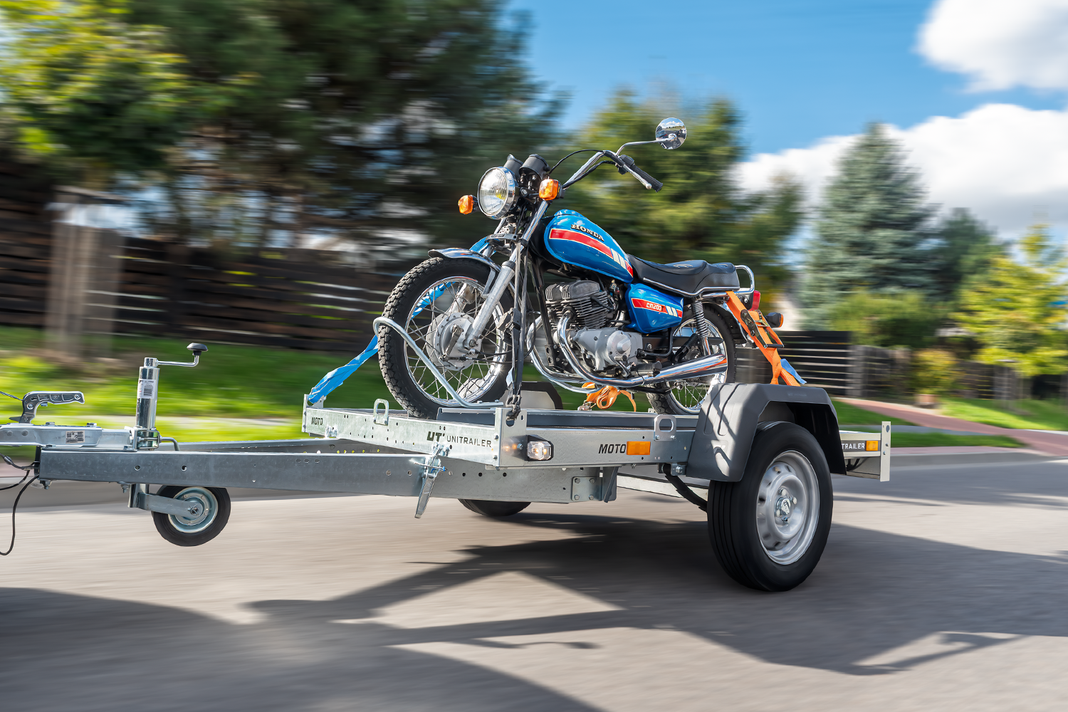 Motocykl typu cruiser zamontowany na płaskiej, srebrnej przyczepie do transportu motocykli. Przyczepka w ruchu, Jasny dzień, w tle pobocze oraz brązowy płot