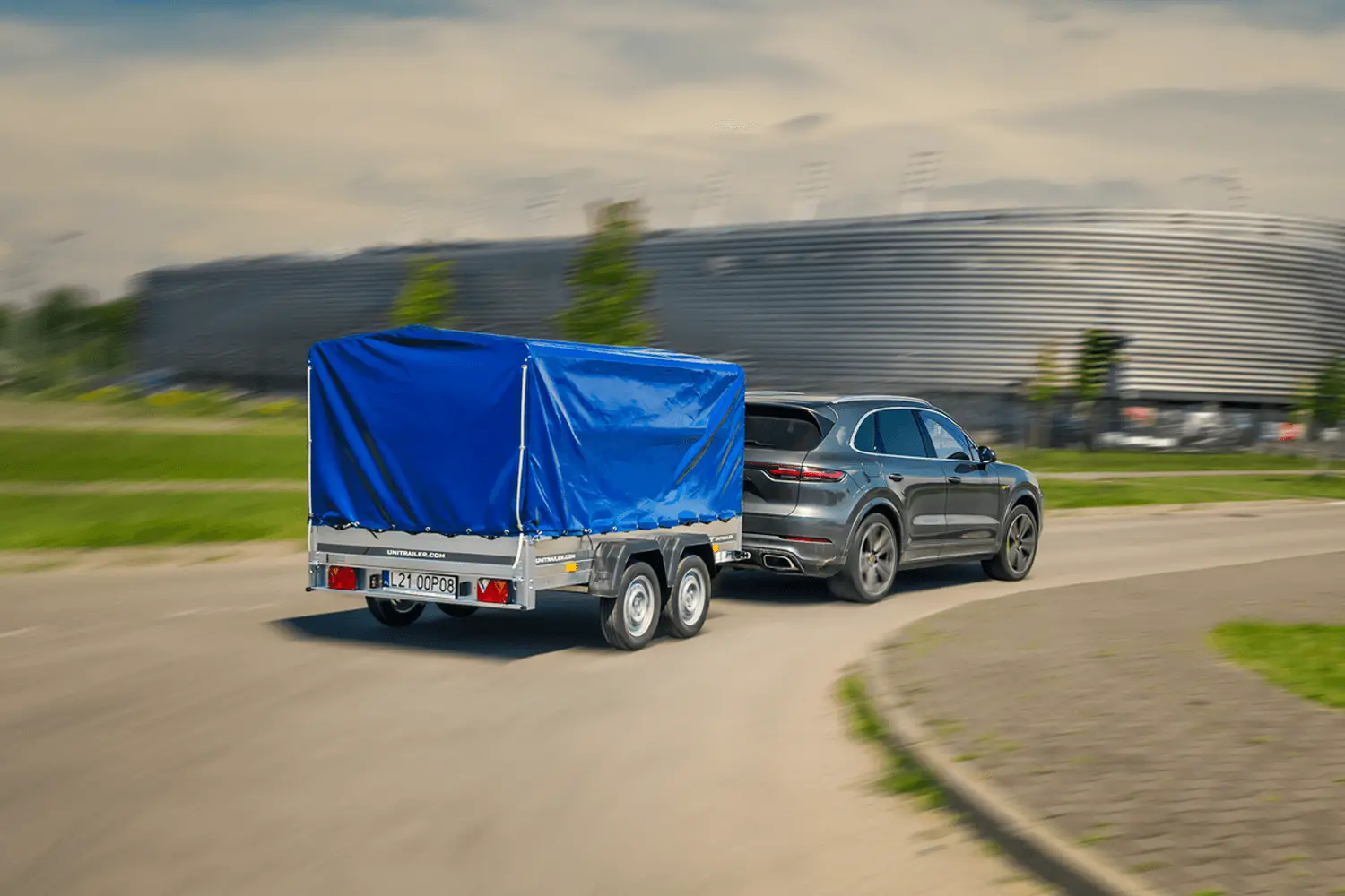Ciemnoszary, nowoczesny SUV (marki Porsche) sfotografowany w ruchu podczas pokonywania łuku na asfaltowej drodze. Pojazd ciągnie stabilną, dwuosiową przyczepę samochodową wyposażoną w wysoki stelaż z naciągniętą, leżącą gładko plandeką w kolorze intensywnie jasnoniebieskim. Na aluminiowej konstrukcji burt oraz na tylnym pasie przyczepy wyraźnie odznacza się adres strony internetowej „UNITRAILER.COM”. Z tyłu zamontowana jest tablica rejestracyjna „L21 0OP08” w otoczeniu czerwonych świateł i trójkątnych odblasków. W tle, za pasem zielonej trawy i młodymi drzewami, widać dużą, nowoczesną bryłę budynku o szarej, metalowej i podłużnej elewacji, przypominającą nowoczesny stadion.