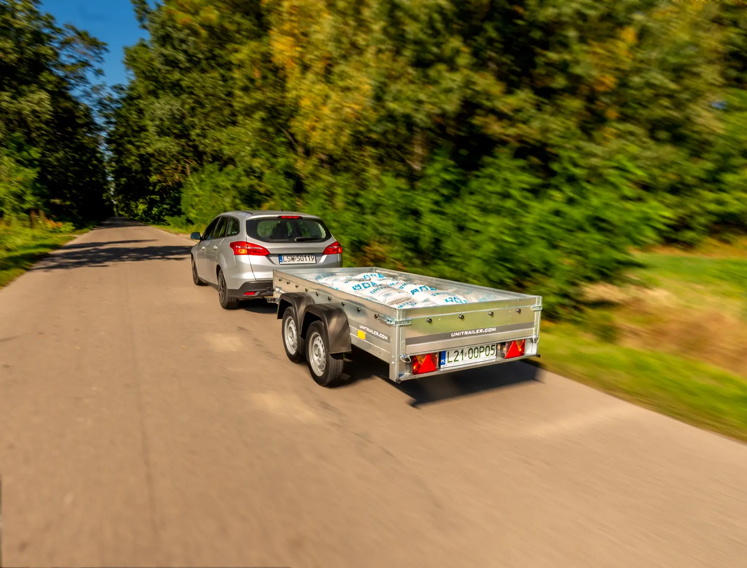 Srebrny samochód osobowy typu kombi (Ford Focus) jedzie lokalną asfaltową drogą, ciągnąc za sobą niską, otwartą przyczepę dwuosiową marki Unitrailer. Przyczepa o odsłoniętej skrzyni ładunkowej jest po brzegi wypełniona ułożonymi płasko białymi workami z niebieskimi nadrukami, w których znajduje się materiał sypki lub budowlany. Na srebrnej, cynkowanej burcie bocznej widnieje napis „UNITRAILER.COM”. Samochód posiada tablicę rejestracyjną „LSW 50119”, natomiast przyczepa „L21 0OP05”. Droga otoczona jest gęstą ścianą lasu z drzewami o zielono-żółtych liściach. Zdjęcie charakteryzuje się dynamicznym rozmyciem tła (panning), co nadaje scenie efekt szybkiego przemieszczania się.