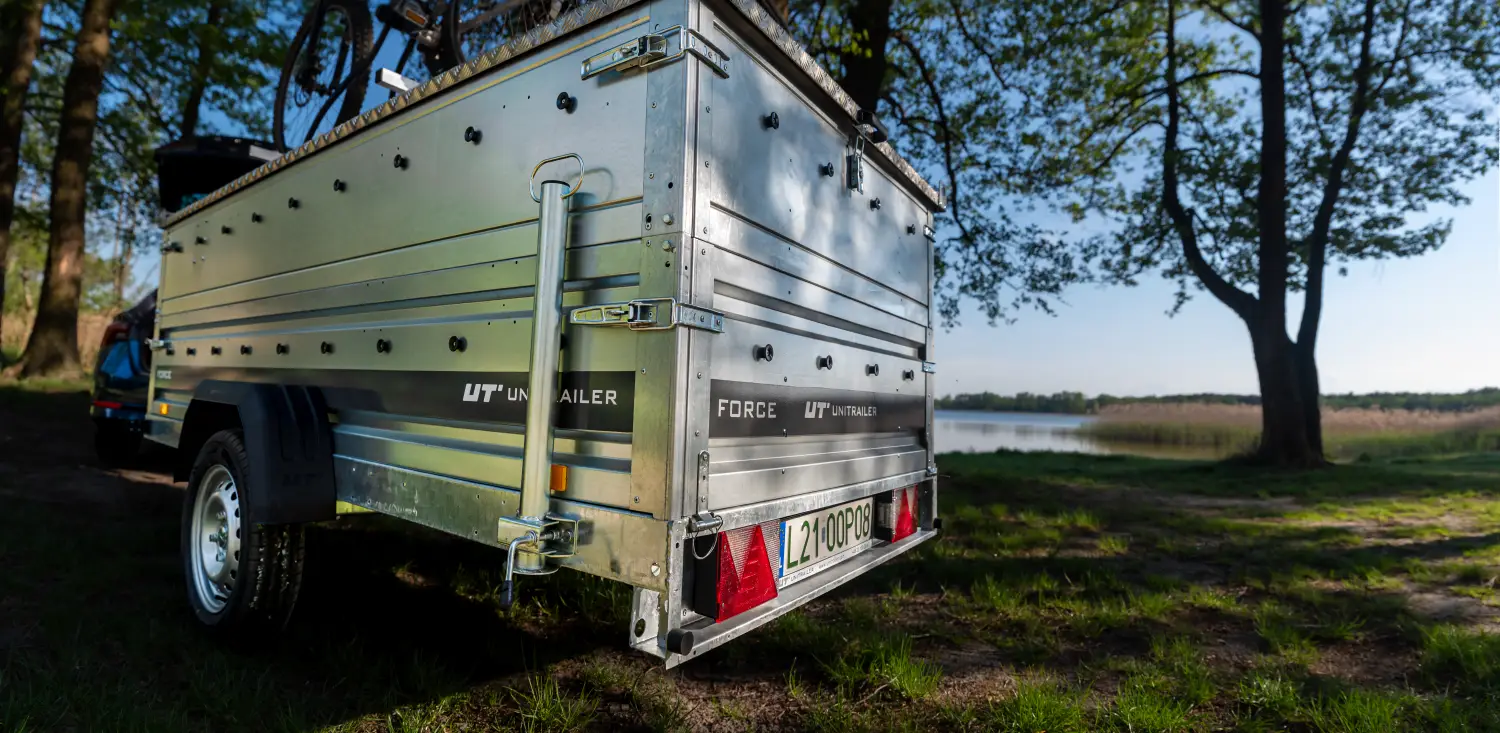Srebrna przyczepa samochodowa Unitrailer FORCE z wysokimi burtami i zamkniętą metalową pokrywą, na której zamontowano rowery, zaparkowana na trawiastym brzegu jeziora w otoczeniu drzew. Z tyłu przyczepy widoczna jest mocowana podpora pionowa z korbą oraz tablica rejestracyjna.