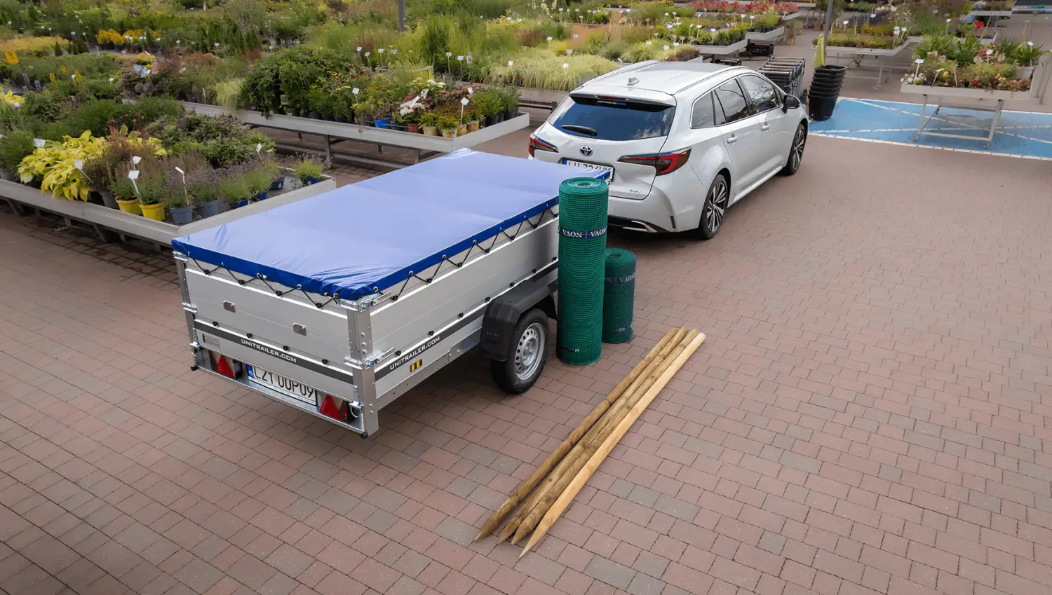 Ujęcie z góry na srebrny samochód kombi z jednonapędową przyczepą Unitrailer zabezpieczoną płaską niebieską plandeką, zaparkowany na kostce brukowej w centrum ogrodniczym obok rolek zielonej siatki i drewnianych palików.