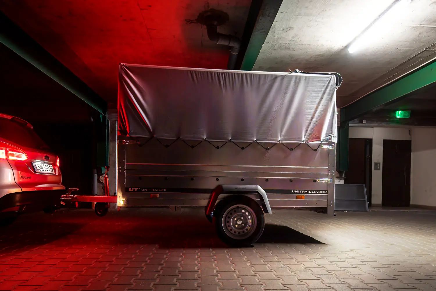 Przyczepa towarowa Unitrailer z szarą plandeką, zapięta do samochodu w słabo oświetlonym garażu podziemnym.