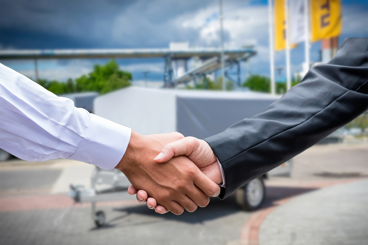 Na pierwszym planie widoczne zbliżenie na uścisk rąk. Widok na ręce od łokcia w dół. Osoba po prawej ubrana w ciemną marynarkę, natomiast po lewej - w jasną koszulę. Tło lekko rozmazane, ale dobrze widoczna jest przyczepka unitrailer z szarym stelażem, żółte flagi unitrailer oraz elementy przemysłowej dzielnicy lublina - sieć grubych, srebrnych rur zamontowana na wielkich konstrukcjach.