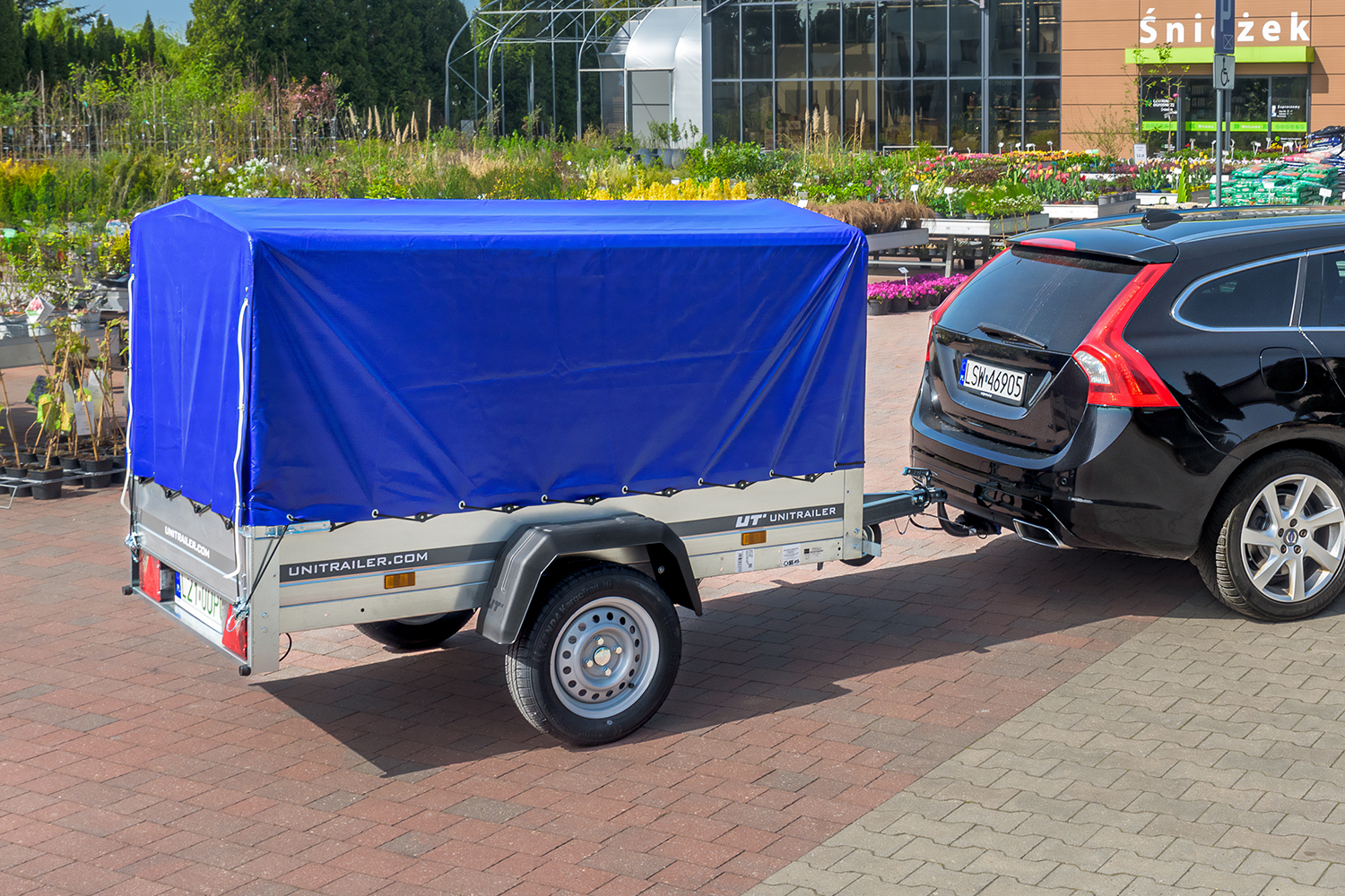 Na pierwszym planie czarne Volvo z podczepioną przyczepką. Przyczepka srebrna z niebieskim stelażem. W tle sklep ogrodniczy 