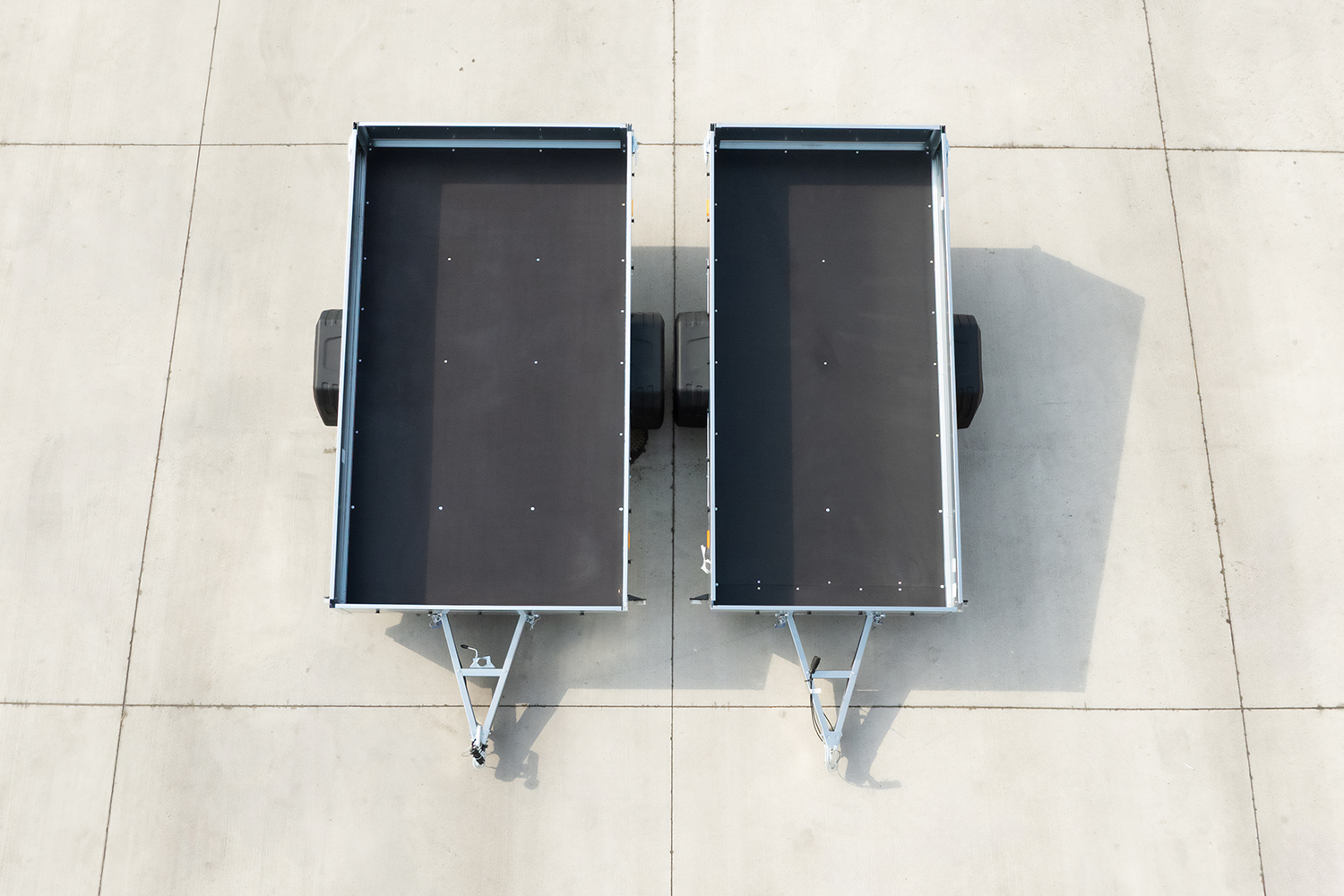 Widok z góry na dwie przyczepki samochodowe ze srebrnymi burtami i czarną podłogą. Jedna szersza, druga węższa. Przyczepki stoją obok siebie, podłoże to duże płyty betonowe.
