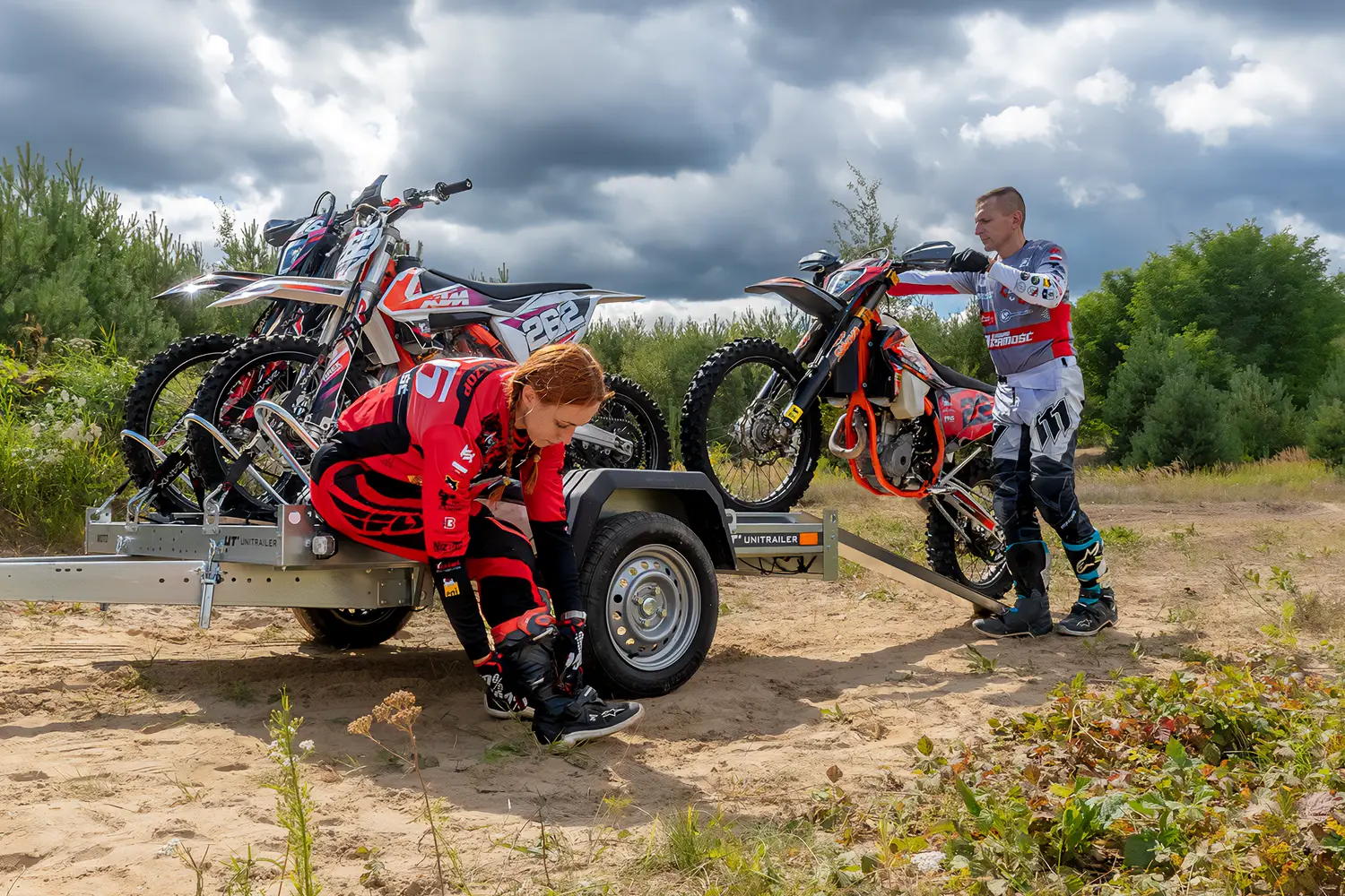 Kobieta i mężczyzna w strojach motocrossowych przygotowują się do jazdy przy przyczepie motocyklowej zaparkowanej na piaszczystym terenie.