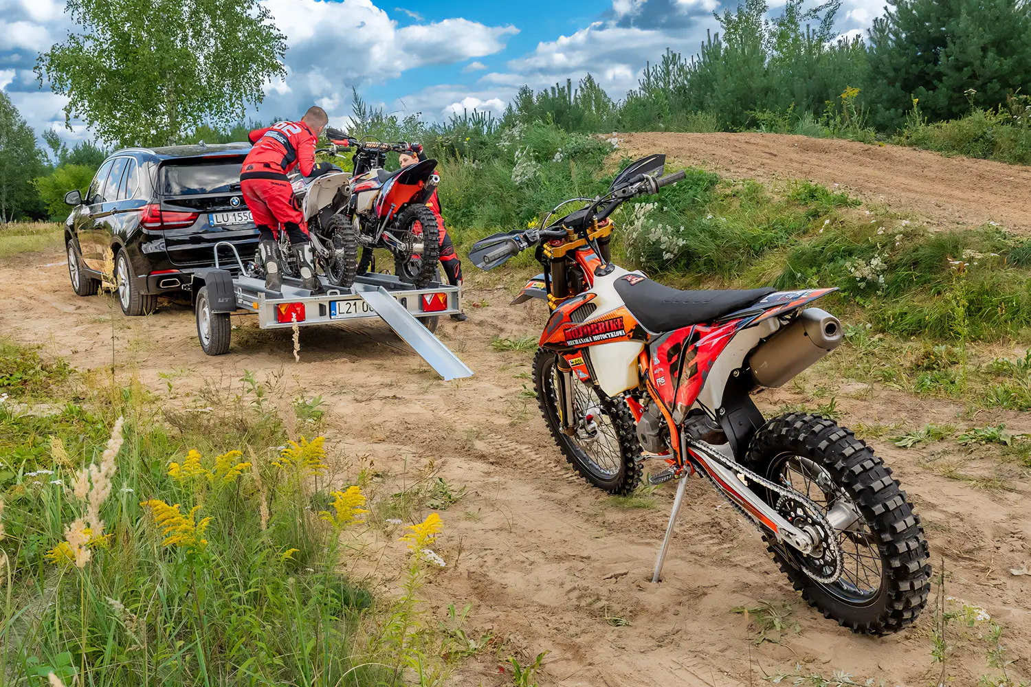 Motocykl crossowy stoi na stopce bocznej na pierwszym planie, a w tle dwie osoby mocują motocykle na przyczepie przypiętej do czarnego SUV-a.