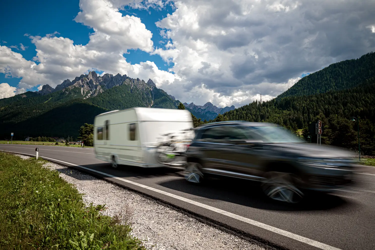 Dynamiczny kadr przedstawiający ciemnoszary samochód typu SUV, który holuje białą przyczepę kempingową na górskiej drodze asfaltowej. Samochód uchwycony w rozmyciu ruchu (motion blur) przemieszcza się szybko w prawą stronę. W tle rozciąga się majestatyczny krajobraz z wysokimi, skalistymi szczytami górskimi pod błękitnym niebem pełnym gęstych, białych i szarych chmur. Po lewej stronie drogi widoczne jest zielone, trawiaste pobocze.