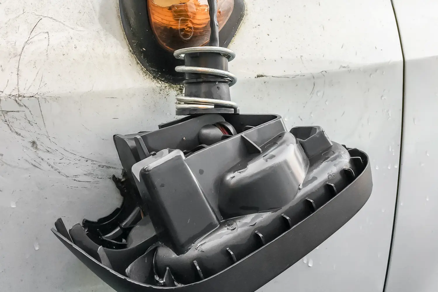 Detal uszkodzenia nadwozia białego samochodu. Czarna, plastikowa obudowa elementu konstrukcyjnego została całkowicie urwana i zwisa do góry nogami na czarnym przewodzie elektrycznym, wokół którego owinięta jest mocna, metalowa sprężyna. Element jest mokry od deszczu, pokryty kropelkami wody i ukazuje wewnętrzne użebrowanie plastiku. Powierzchnia białego lakieru wokół urwanego mocowania oraz podłużnego, pomarańczowego klosza lampy jest wyraźnie porysowana i zabrudzona.