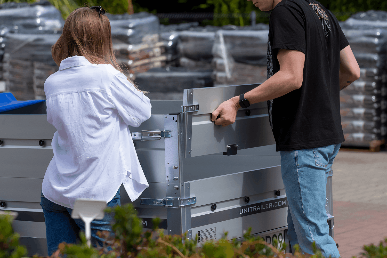 Bliski kadr pokazujący dwie osoby obsługujące tylną burtę przyczepy samochodowej marki Unitrailer. Mężczyzna trzyma za uchwyt dodatkowej, aluminiowej nadstawki burty, składając ją lub montując. Obok stoi kobieta w białej koszuli i okularach przeciwsłonecznych na głowie. Na zdjęciu wyraźnie widać detale konstrukcyjne przyczepy: srebrne aluminiowe burty, ocynkowane zamki dźwigniowe burt, czarne plastikowe uchwyty do mocowania linki plandeki oraz napis UNITRAILER.COM.