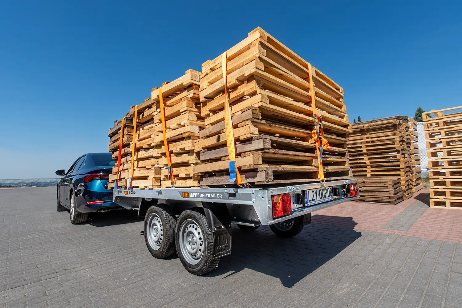 Dwuosiowa przyczepka towarowa Unitrailer spięta z ciemnym samochodem, załadowana wysoką stosem drewnianych palet zabezpieczonych pasami transportowymi na placu firmowym.