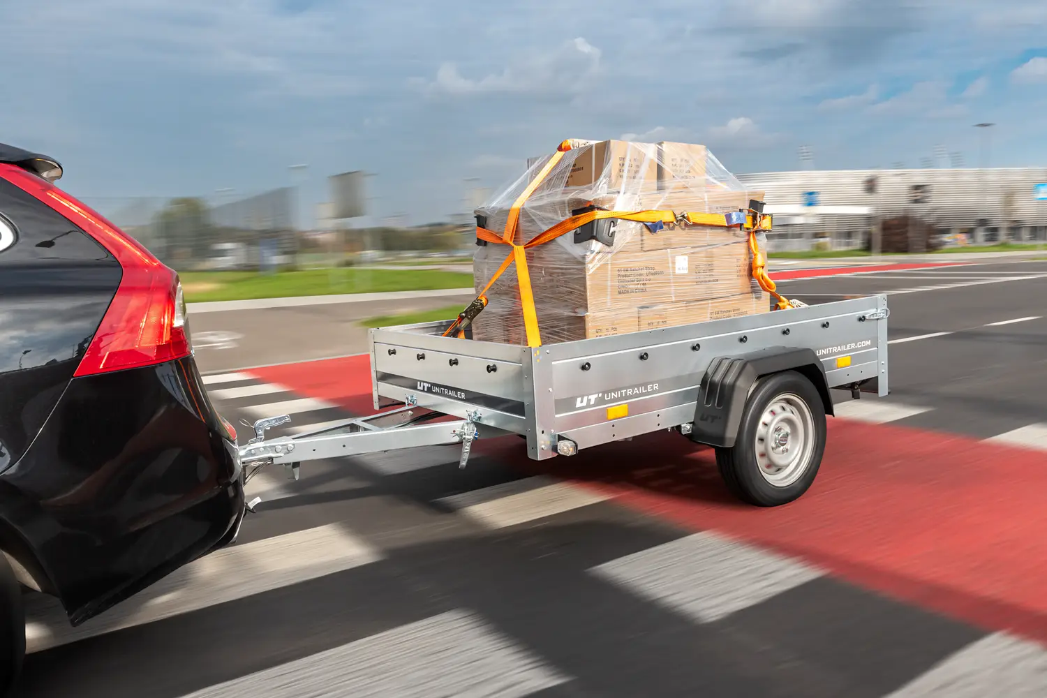 Przyczepka towarowa Unitrailer spięta z czarnym samochodem, przewożąca paletę z kartonami owiniętą folią stretch i zabezpieczoną pomarańczowymi pasami transportowymi podczas jazdy po drodze